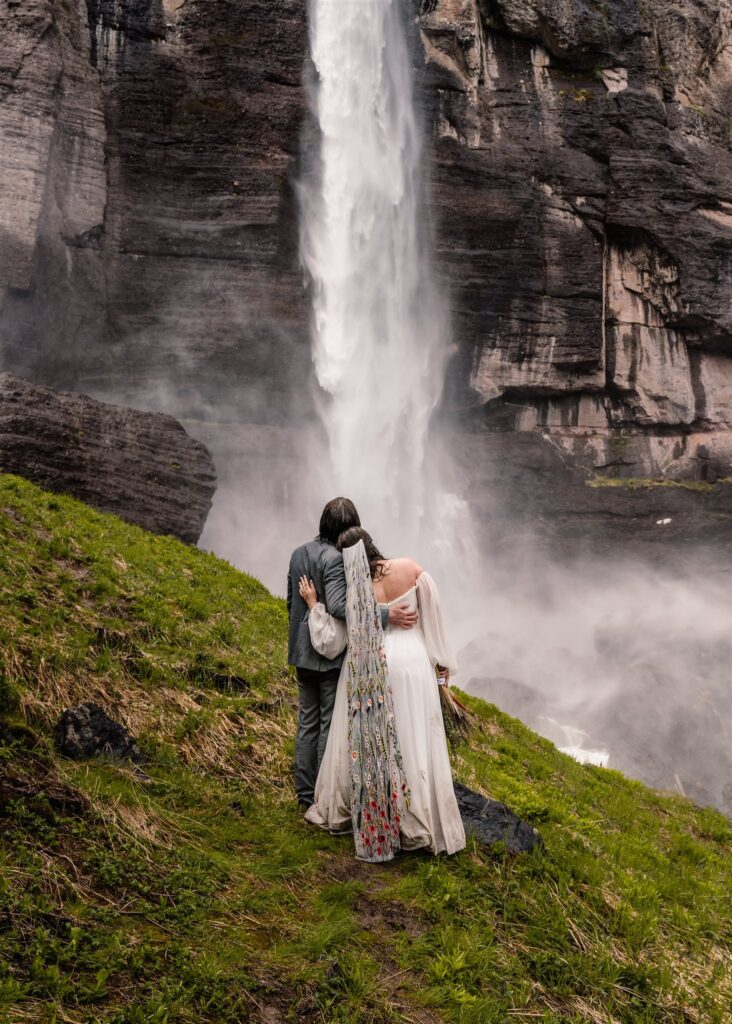 A bride and groom in their wedding attire gaze out at a gushing waterfall after their sunrise elopement