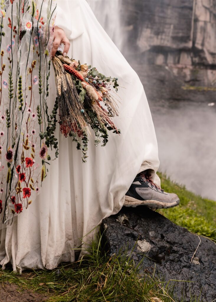 A vertical shot focused on a dried, bridal bouquet, the end of a veil embroidered with colorful wildflowers, and muddy hiking boots.  