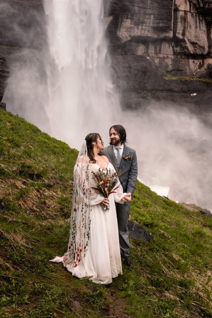 A bride and groom stand in front of the gushing falls and gaze at each other after their sunrise elopement