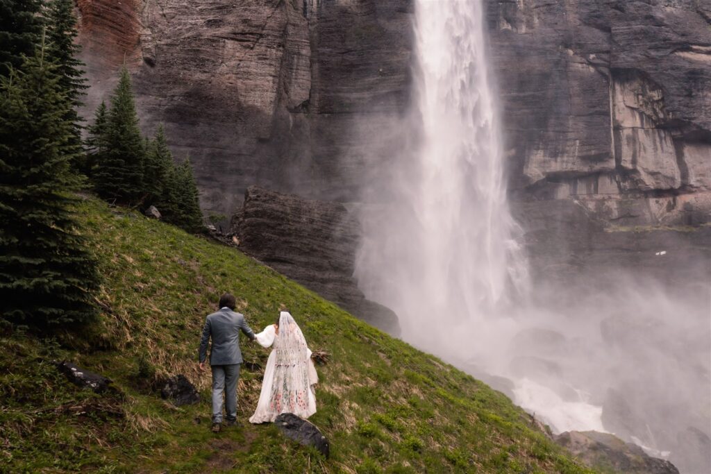 A wide shot of a couple in wedding attire walking a long a green trail. They walk hand in hand towards the gushing falls during their sunrise elopement