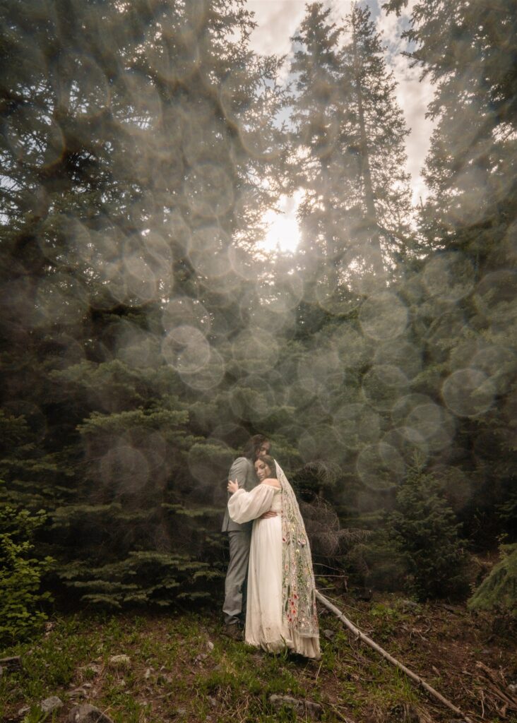 A bride and groom in wedding attire embrace during their sunrise elopement. Dew dapples the camera lens and creates almost a disco ball affect