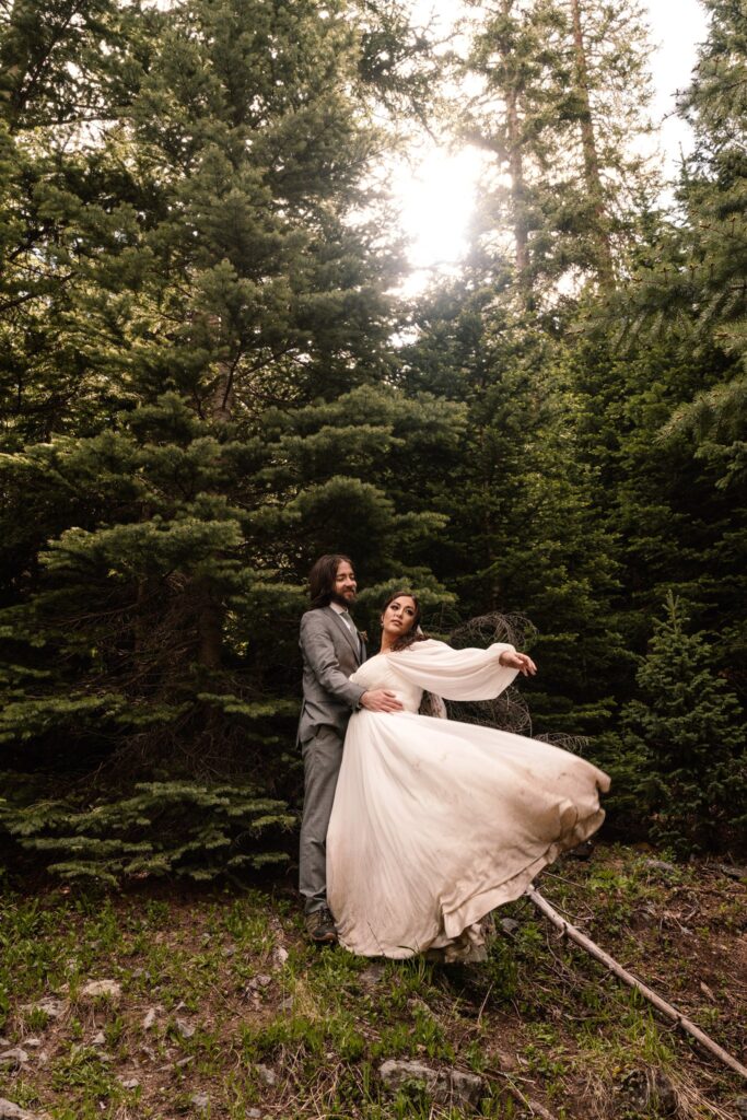 golden light filters through the treetops during this sunrise elopement. A bride and groom embrace as the brides dress billows behind her