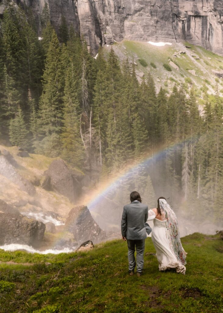 A bride and groom in their wedding attire walk towards a rainbow during their sunrise elopement