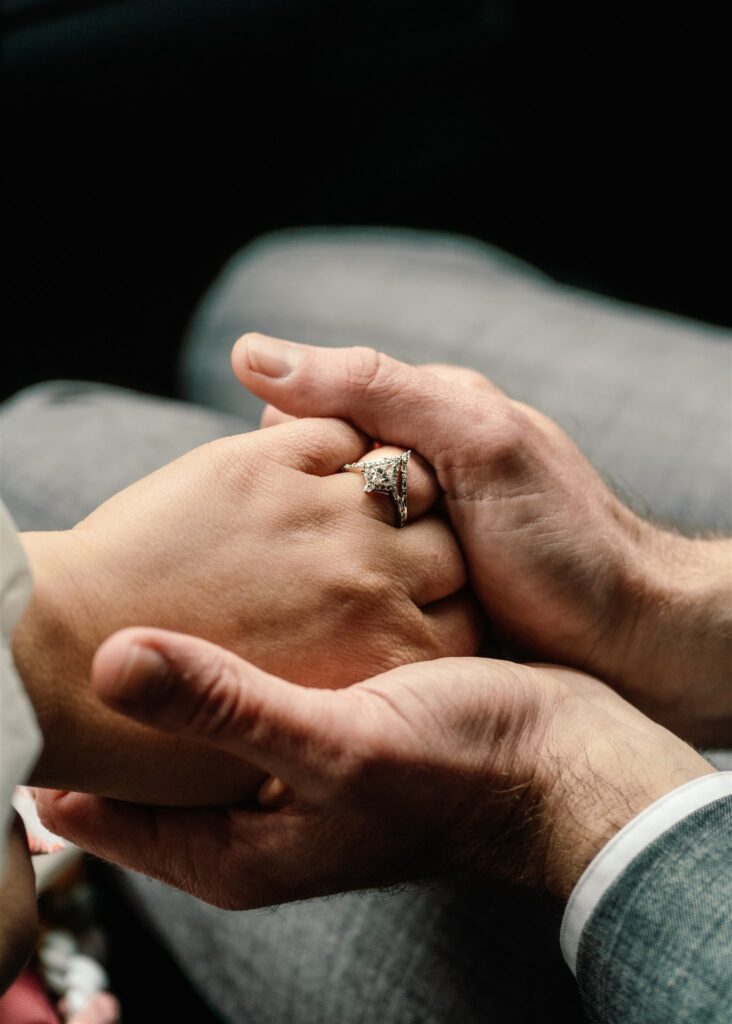 A close up image of a  bride and groom holding hands. the focus is on the brides ring finger where we see her newly places wedding band. 