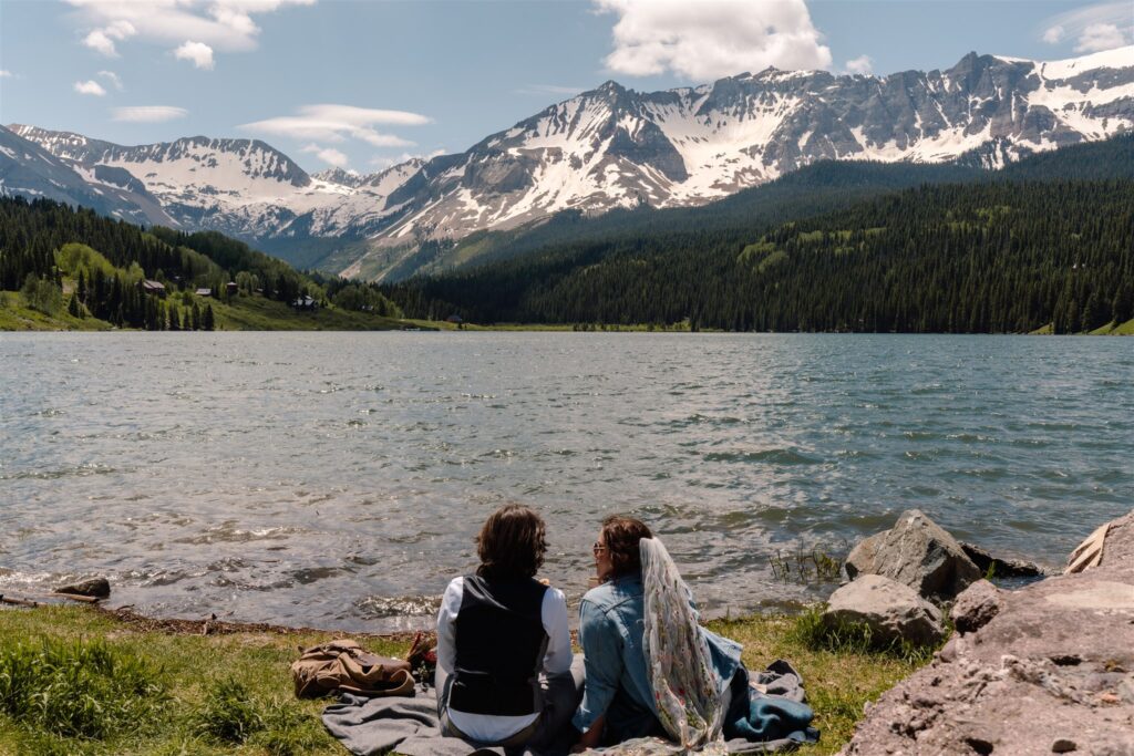 Mountains surrounding them, a couple in wedding attire sit on the shore of a gorgeous, blue mountain lake, celebrating after their sunrise elopement.