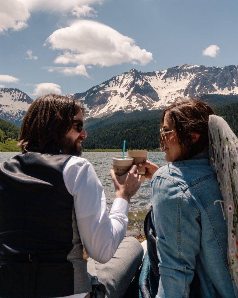 a couple in wedding attire clink their iced coffee on the shores of a mountain lake after their sunrise elopement