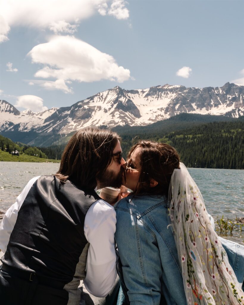 A bride and groom kiss in front of a beautiful, blue lake, surrounded by mountains. They are in their wedding attire, but have added casual elements like a jean jacket and sunglasses for a more dressed-down look for after their sunrise elopement