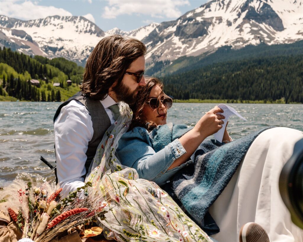 A bride and groom lay on the shore of a beautiful blue lake. The snowy mountains rise above them as they recline and read letters from friends and family after their sunrise elopement