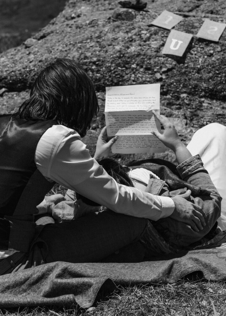 A black and white photo of a bride and groom reading letters from family after their sunrise elopement. Groom sits on a blanket while his bride lays her head in his lap