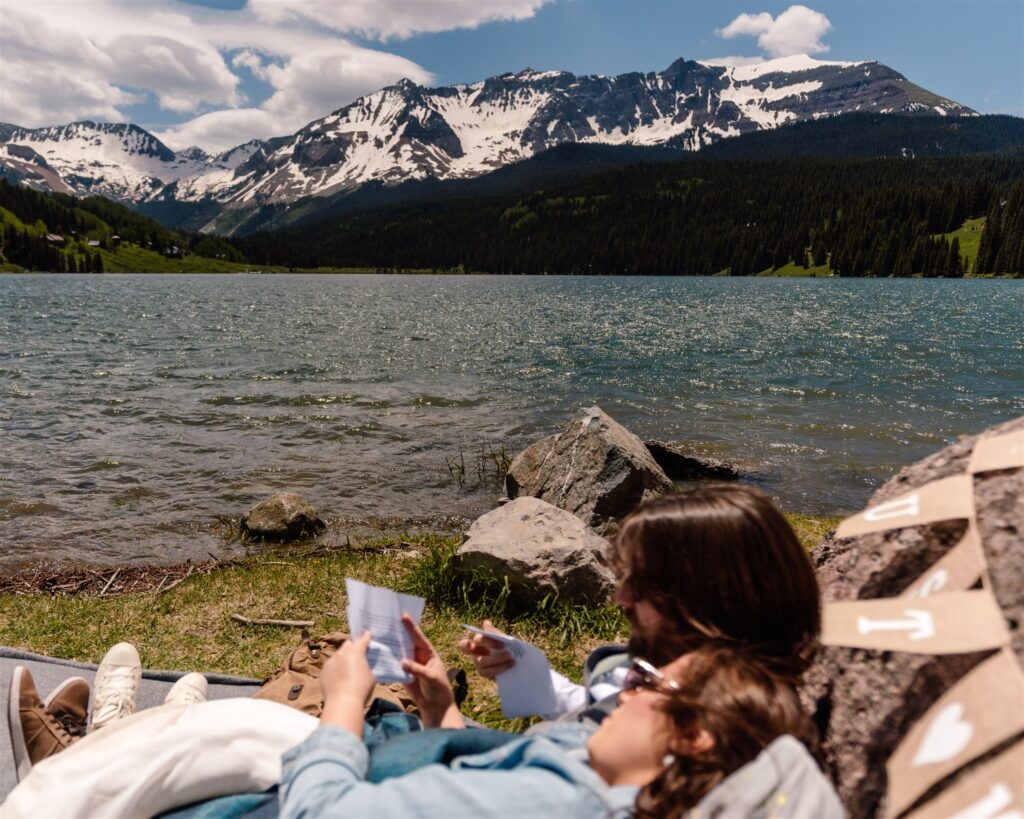 A bride and groom lay on the shore of a beautiful blue lake. The snowy mountains rise above them as they recline and read letters from friends and family after their sunrise elopement