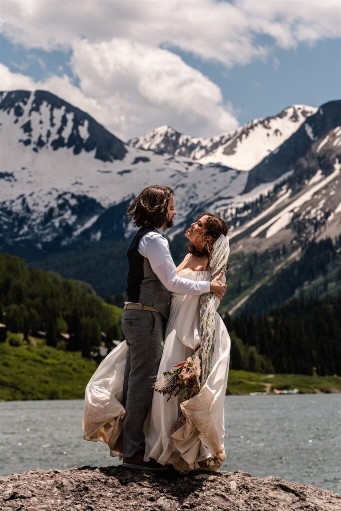 the wind whips a couple in wedding attire as they embrace on a large rock after their sunrise elopement. behind them is a gorgeous lake surrounded by mountians