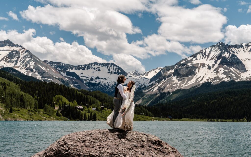 a couple in wedding attire embrace while standing on a large rock. Behind them is a gorgeous, blue lake surrounded by snow-capped mountains.