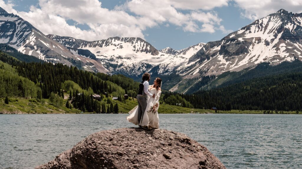 A bride and groom embrace on a large rock in their wedding attire. The wind wwhips the brides dress and they gaze at each other during their sunrise elopement