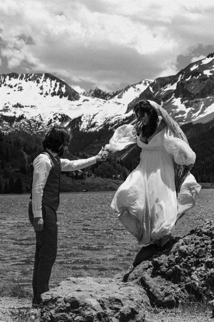 A black and white photo of a groom helping his bride down from a large rock. mountains and a beautiful lake surround them after their sunrise eloepment