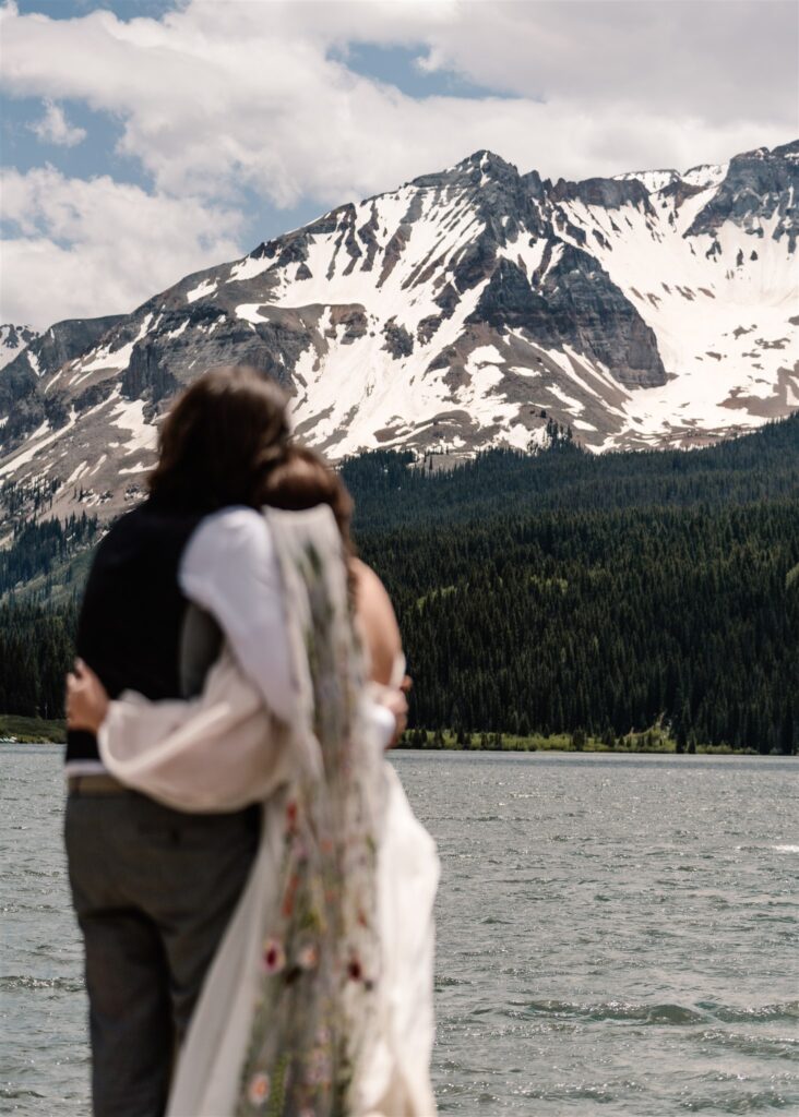 a shot of snow-capped mountians that tower above a beautiful blue lake. A couple in wedding attire embrace in front of the lake after their sunrise elopement