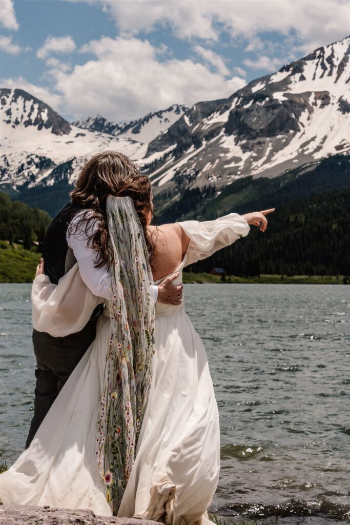 a shot of snow-capped mountains that tower above a beautiful blue lake. A couple in wedding attire embrace in front of the lake after their sunrise elopement. The bride points to something in the distance.