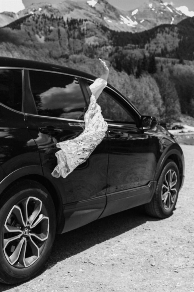 A black and white photo of a car driving off. from the passenger window, we see a bridal veil billowing in the wind as she waves, ending their sunrise elopement