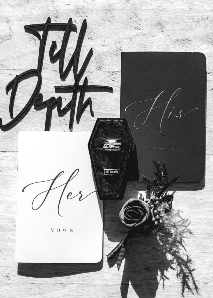 a black and white image of this moody pacific northwest elopement wedding details including a wooden sign that reads "til death", his and her vow books, a boutonnière, and wedding rings resting in a velvet coffin ring box 