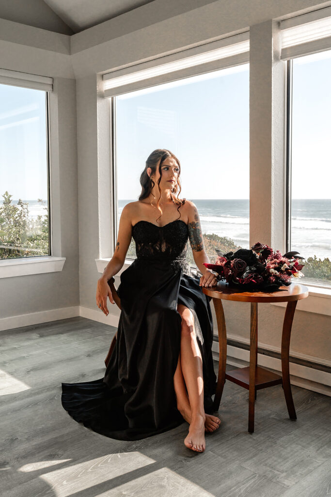 a bride in an all black wedding dress sits beside a table with her bouquet resting beside her. behind here are large windows that show thee ocean below them  