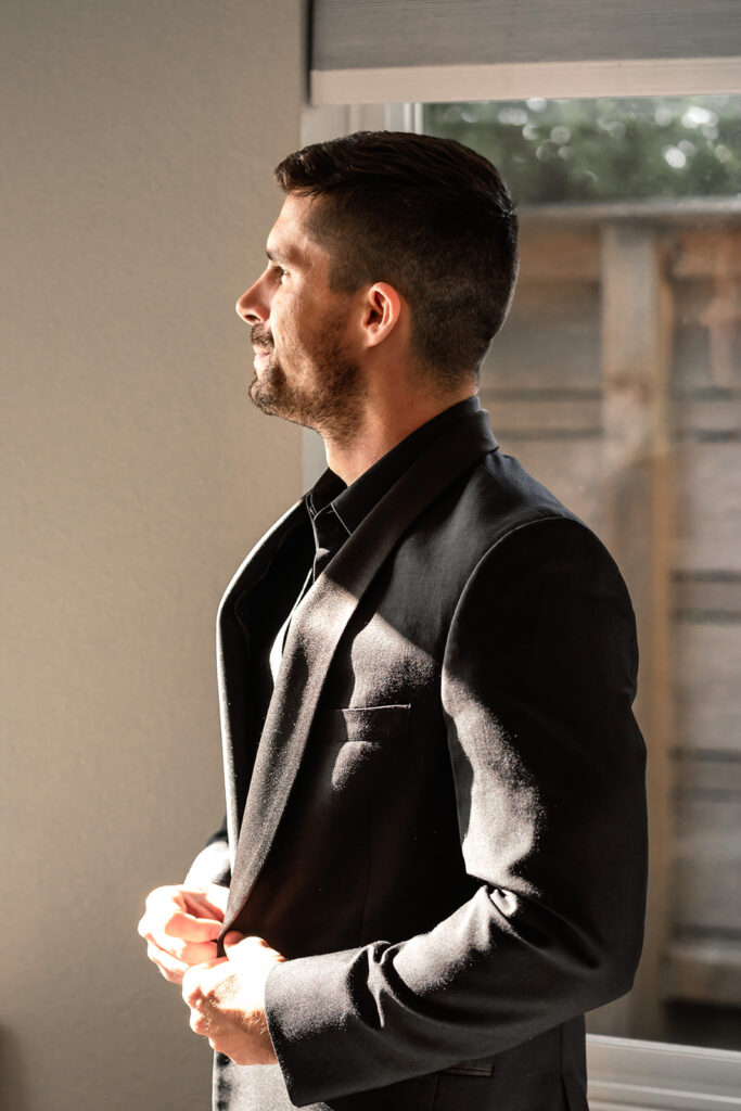 a groom in an all black suit gazing out the window after getting ready for his pacific northwest elopement