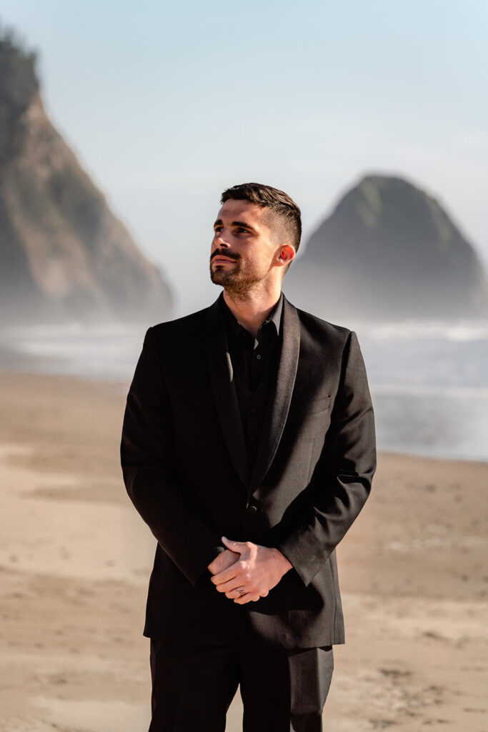 a groom stands on a rocky beach in all black wedding attire. The sun shines brightly on him as he gazes towards his bride