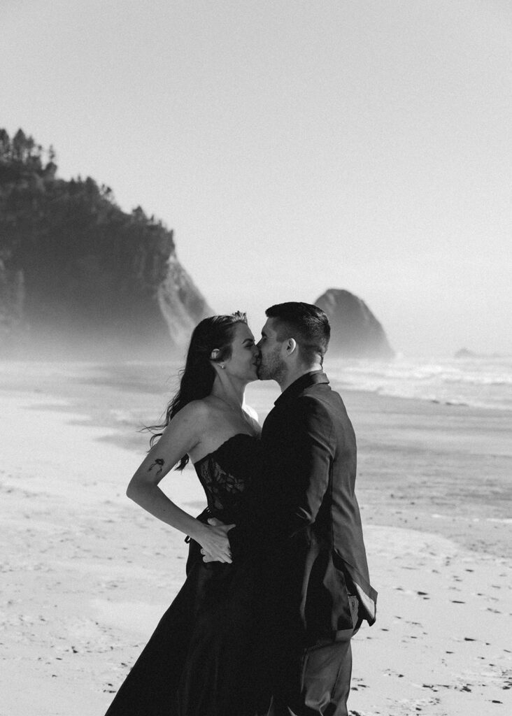 a black and white photo of a bride and groom in all black wedding attire kissing on a rugged beach during their Pacific northwest elopement