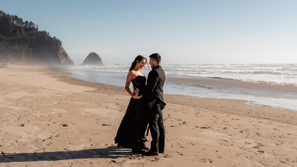 a bride and groom in all black wedding attire embrace in a rocky beach. The sun shines brightly on them as they hug during their pacific northwest elopement