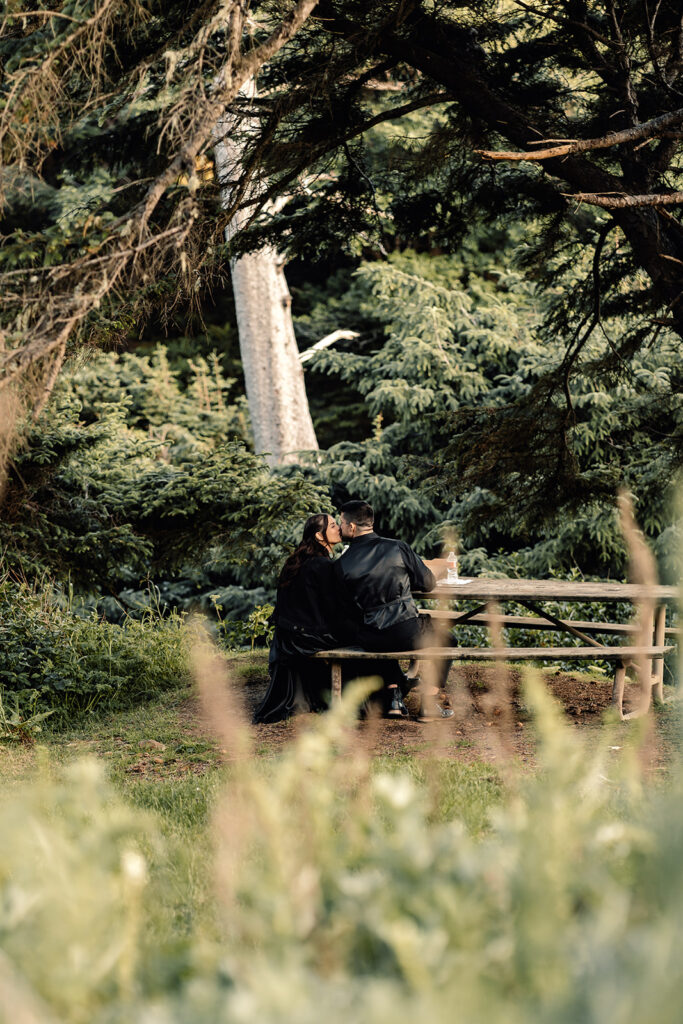 a shot of a couple in all black wedding attire kissing while sitting at a picnic table during their pacific northwest elopement