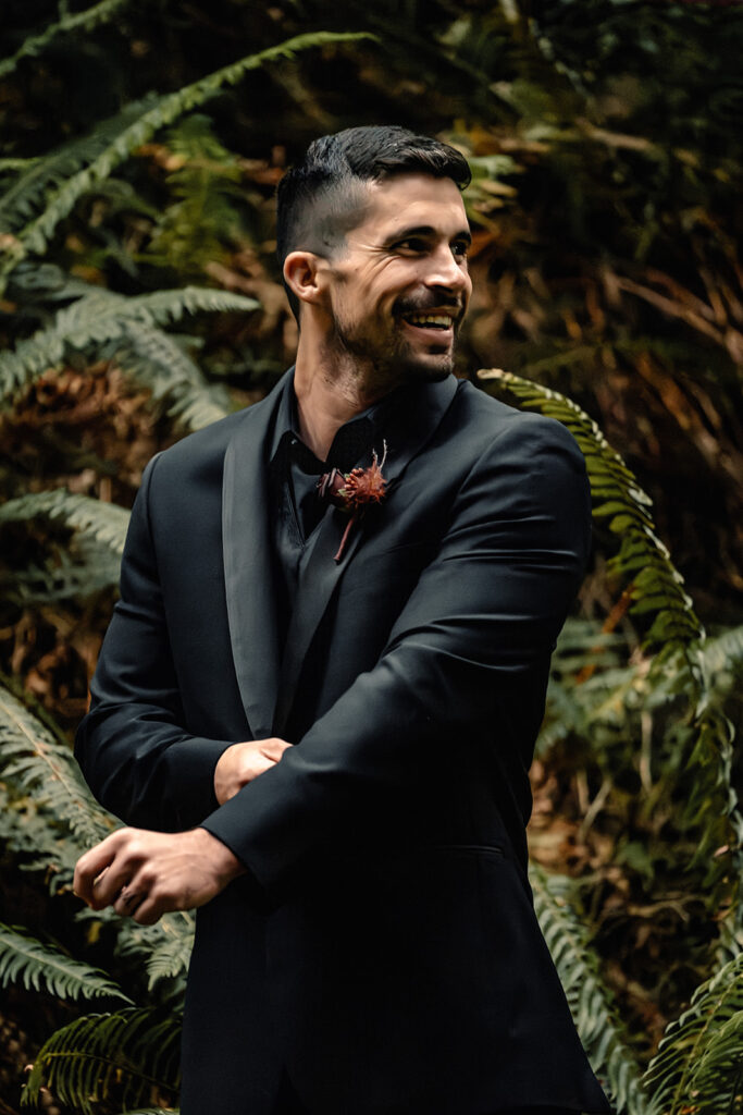 a groom stands surrounded by ferns in an all black suit. he smiles widely as he smiles back at his bride