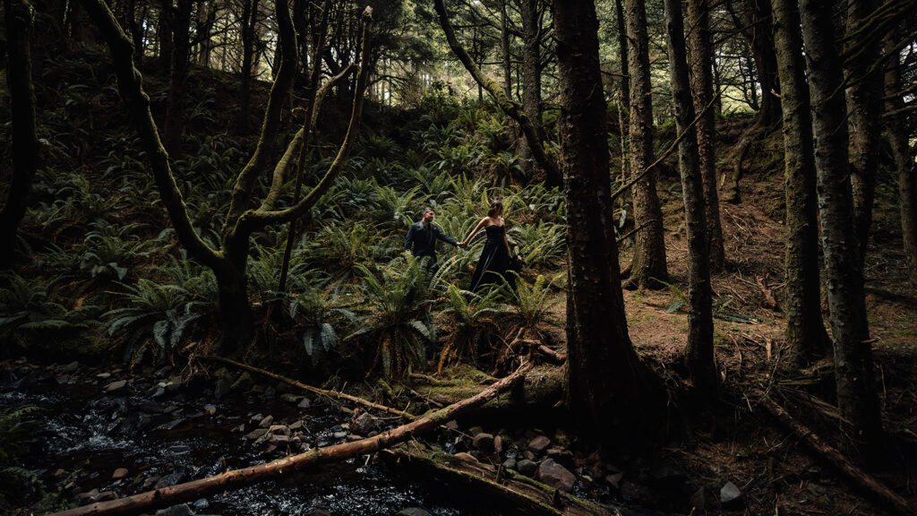 a bride and groom in all black wedding attire explore a moody forest full of lush ferns and mossy trees during their pacific northwest elopement