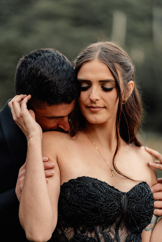 A couple in all black wedding attire embrace. The groom kisses his brides shoulder as she caresses his face during their pacific northwest elopement 