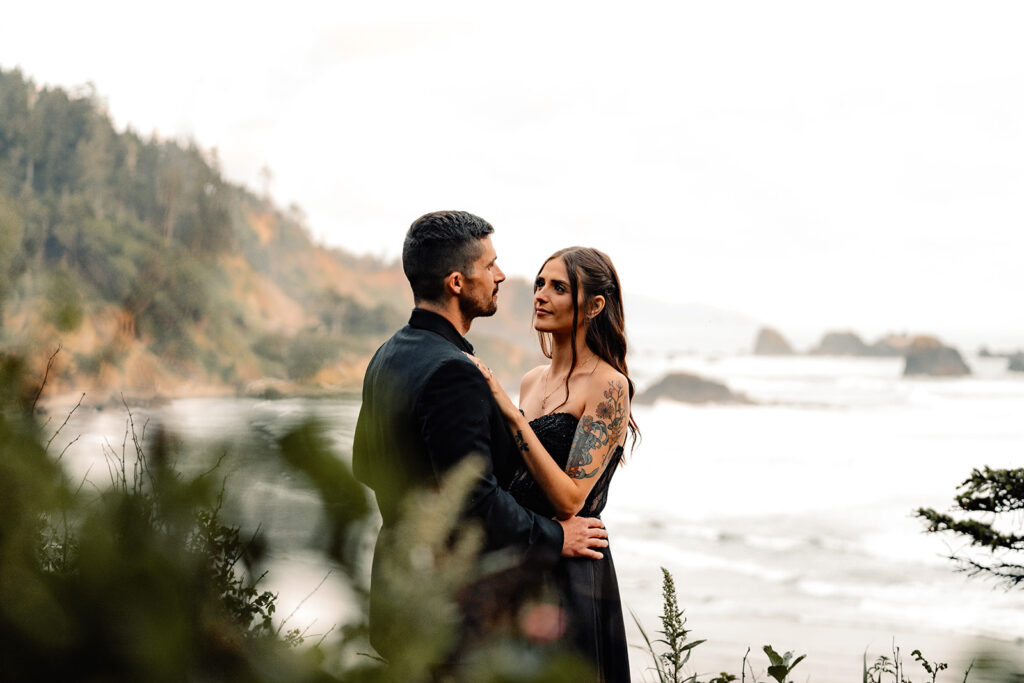 A couple in all black wedding attire embrace on the edge of an ocean cliff during their pacific northwest elopement. they are framed by the surrounding greenery