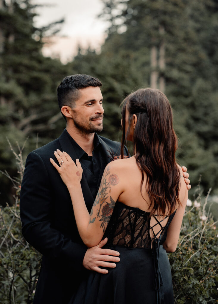 A couple in all black wedding attire embrace. The groom smiles sweetly at his bride during their pacific northwest elopement 