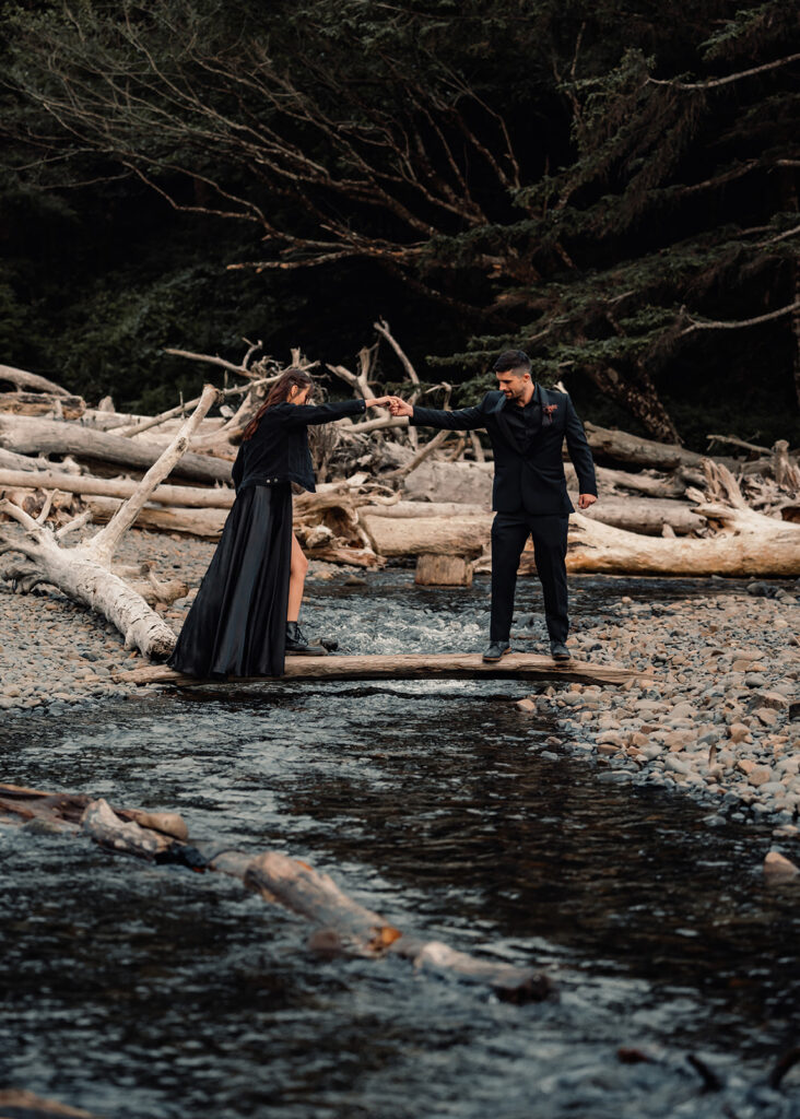 a groom helps his bride cross a small stream. they both wear all black wedding attire during their pacific northwest elopement