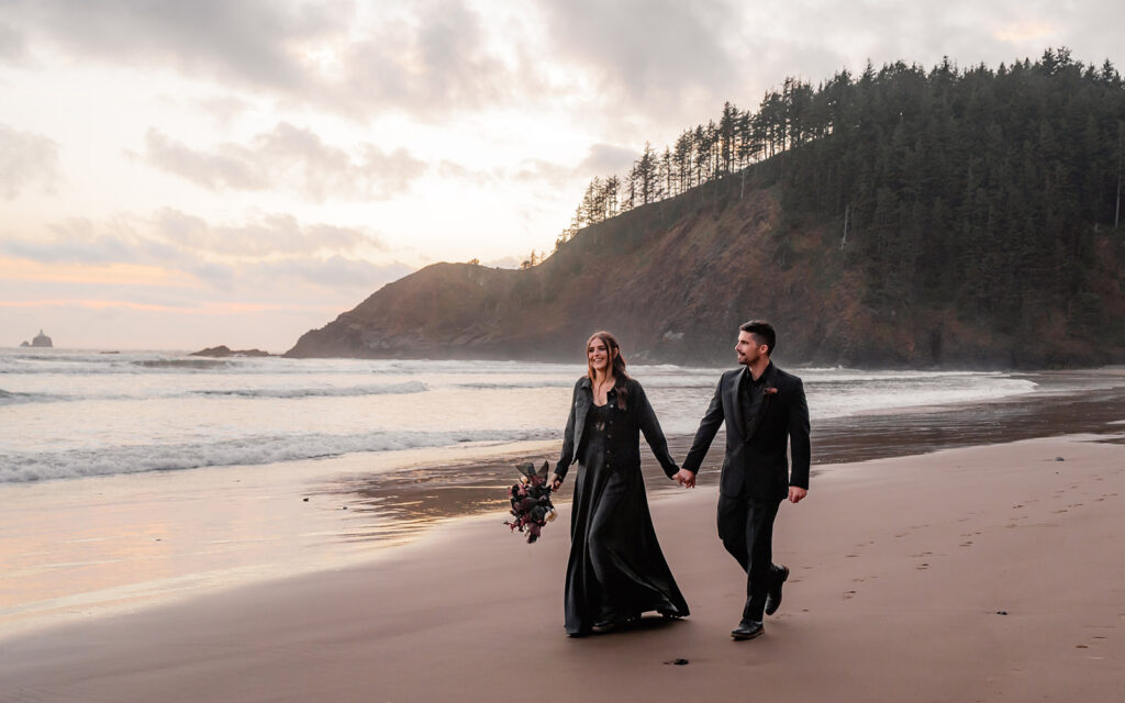 a bride and groom in all black wedding attire explore a rocky beach hand in hand during their pacific northwest elopement