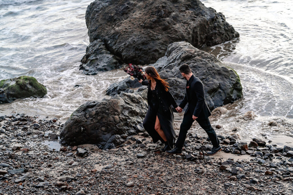 a bride and groom in all black wedding attire explore a rocky beach hand in hand during their pacific northwest elopement