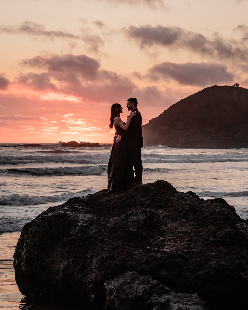 a couple in all black wedding attire embrace on top of a rock during the sunset of their pacific northwest elopement