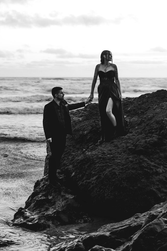 a couple in all black wedding attire stand hand and hand on top of a rock during the sunset of their pacific northwest elopement