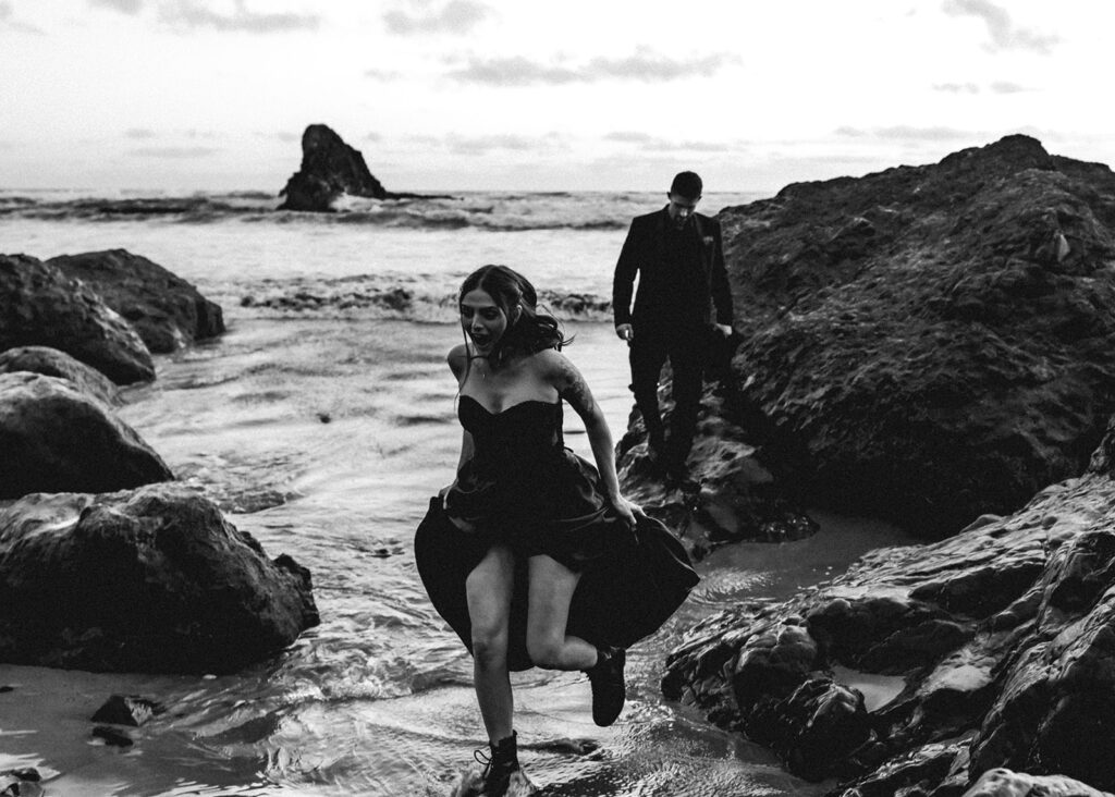 a couple in all black wedding attire descend from a large rock and run from the incoming waves during their pacific northwest elopement