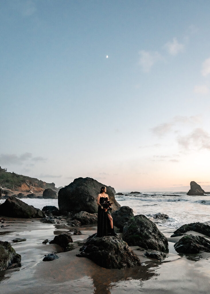 a bride in all black wedding attire stands on a jagged rock, the glowing moon hangs directly above her during her pacific northwest elopement