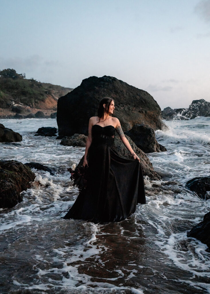 a bride in all black wedding attire stands on a jagged rock, the waves swirl around her during her pacific northwest elopement