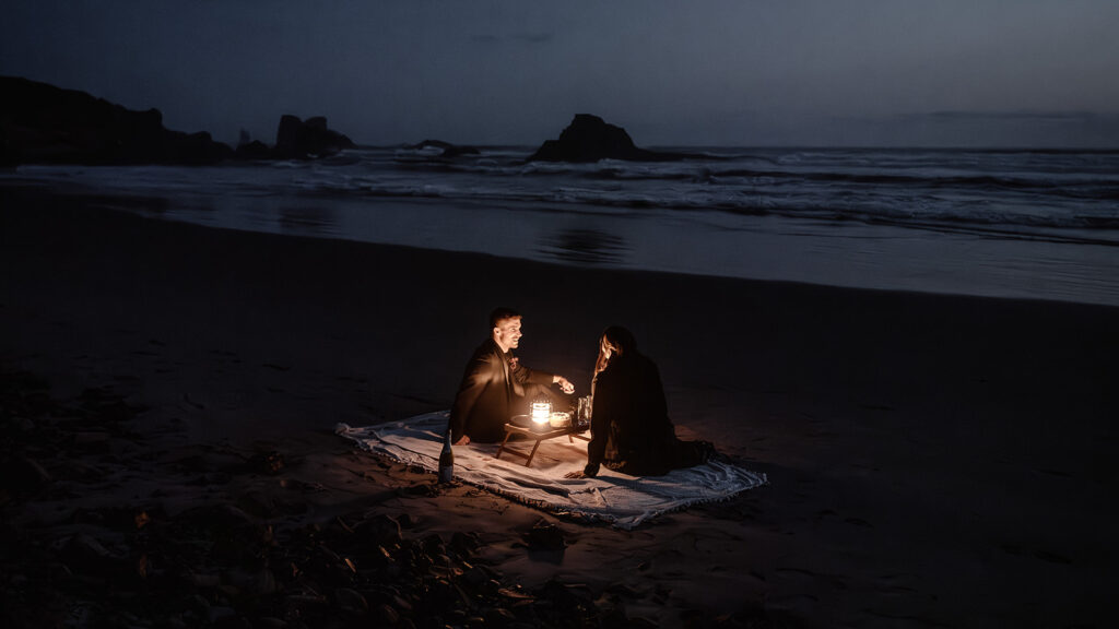 a couple in all black wedding attire share a lantern lit picnic on the beach during their pacific northwest elopement. the shadows of the sea stacks can be seen in the distance