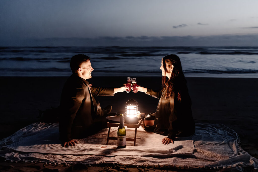 a couple in all black wedding attire share a lantern lit picnic on the beach during their pacific northwest elopement. They toast their drinks together in celebration