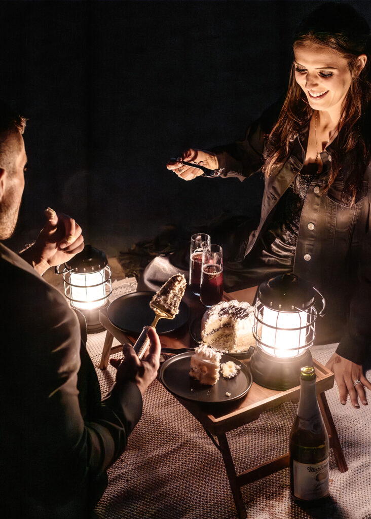 a couple in all black wedding attire share a lantern lit picnic on the beach during their pacific northwest elopement. there is a cake that they share as they smile