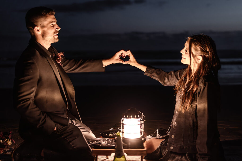 a couple in all black wedding attire share a lantern lit picnic on the beach during their pacific northwest elopement. the connect their hands over their picnic table too create a heart shape