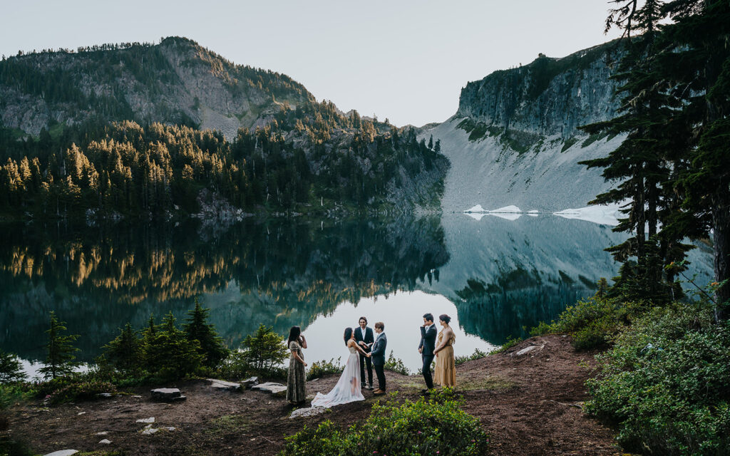 a bride and groom in their wedding attire exchange vows on the shores of an alpine lake during their hiking elopement. they are surrounded by a small group of good friends