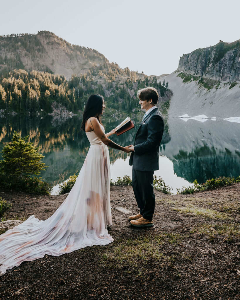 a bride and groom in their wedding attire exchange vows on the shores of an alpine lake during their hiking elopement