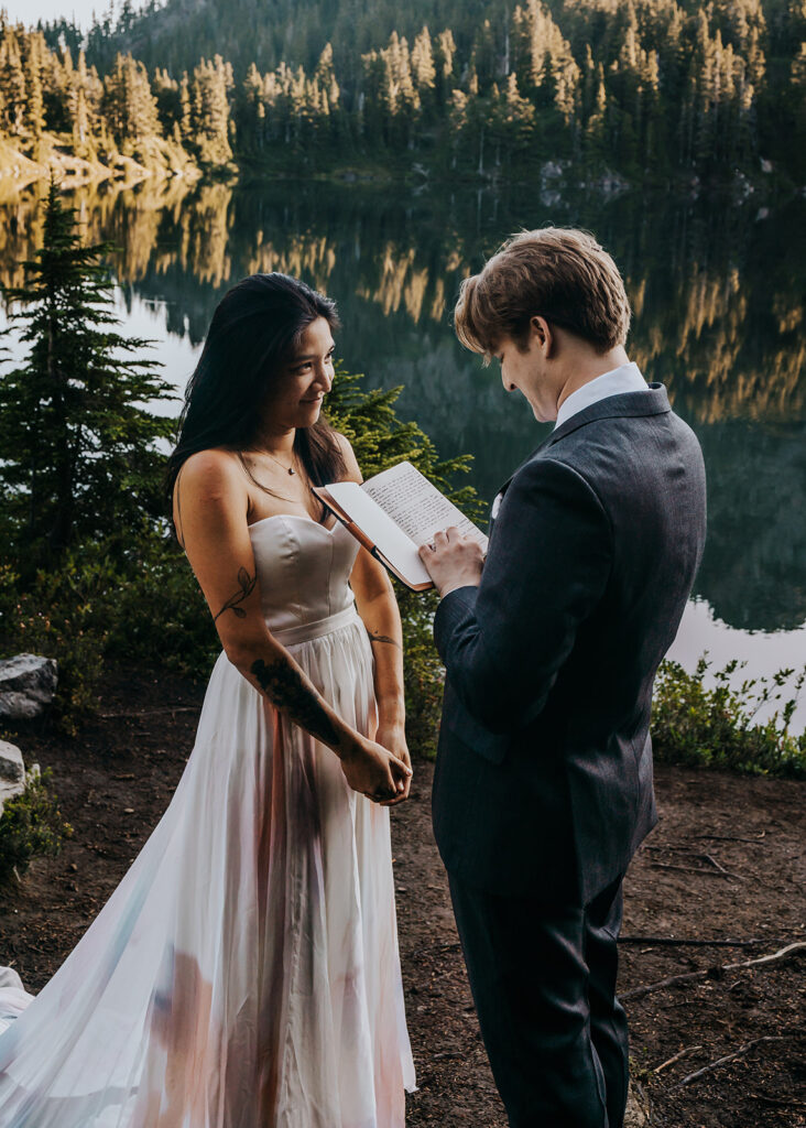 a bride and groom in their wedding attire exchange vows on the shores of an alpine lake during their hiking elopement