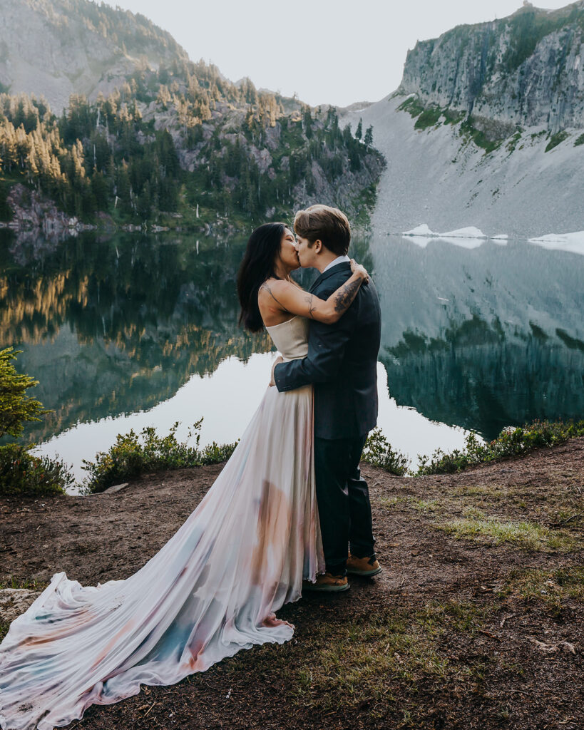 a bride and groom in their wedding attire share their first kiss on the shores of an alpine lake during their hiking elopement