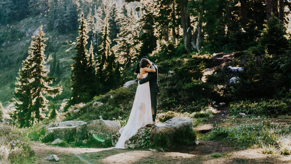 a couple in their wedding attire, embrace while standing on a large rock. In the background, we see a beautiful lake and a mountain, setting the scene for their hiking elopement.